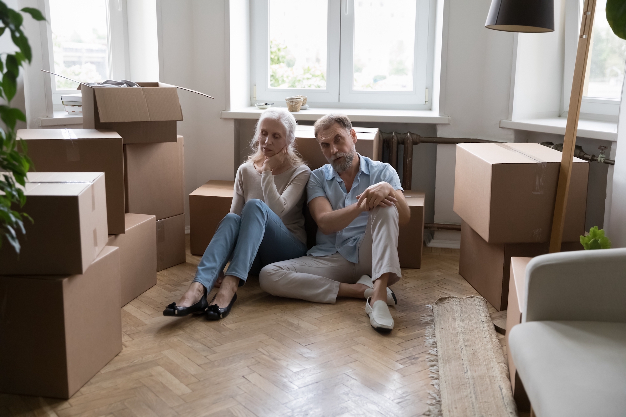 Older couple sit in a dark room with lots of moving boxes around and look depressed